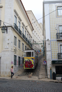 Graffiti-covered tram traveling uphill on a narrow, steep street between tiled buildings in Lisbon, Portugal.