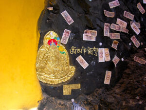 Rock surface painted with a golden religious figure, Tibetan script, and attached paper offerings, photographed inside a cave in Tibet.