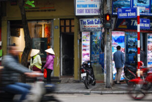 Busy urban street with pedestrians, a parked motorbike, and signage including “The Sinh Café,” photographed in Hanoi, Vietnam.